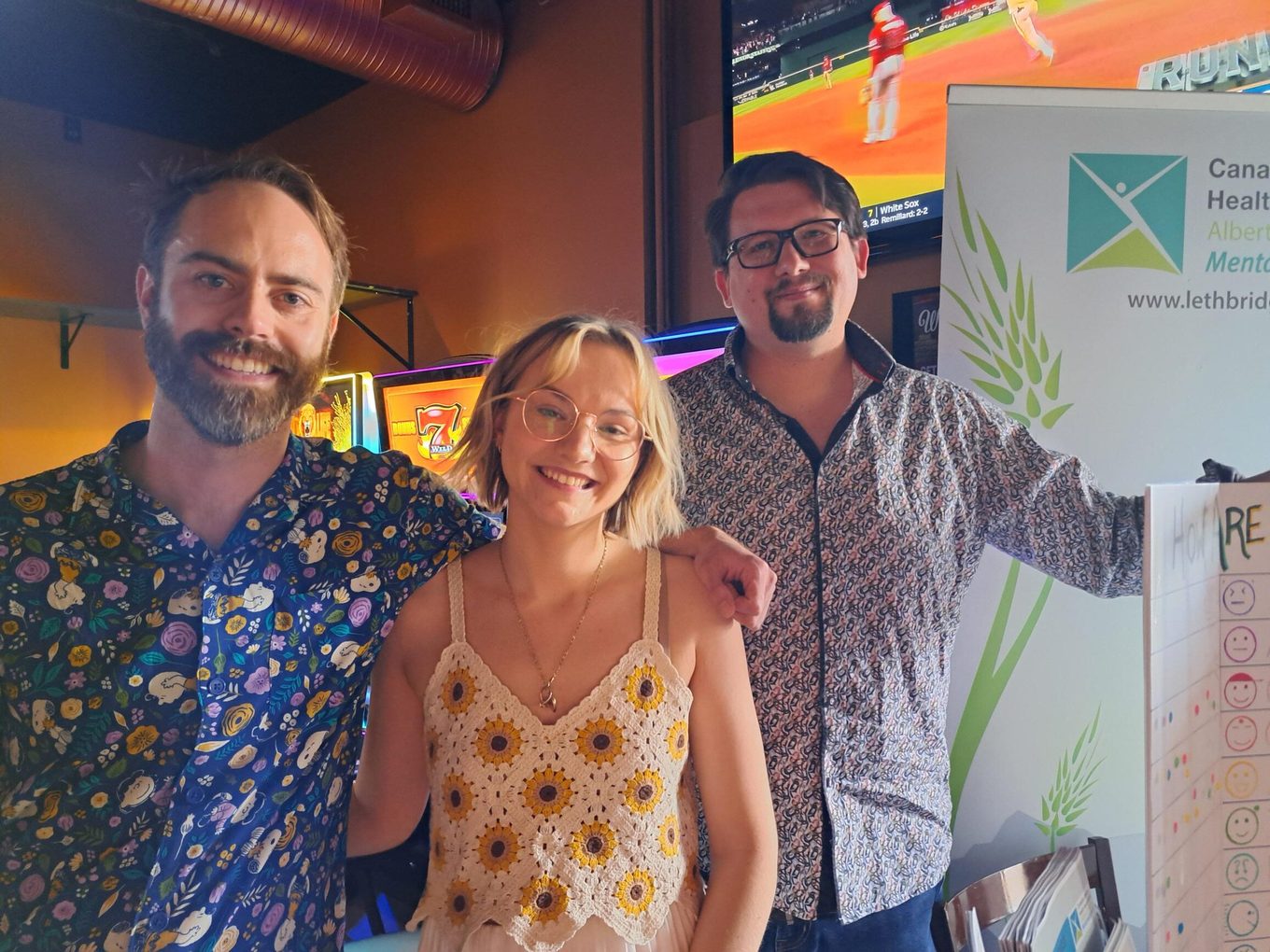 Three people standing shoulder to shoulder and smiling inside a pub, in front of a pull-up banner for the Canadian Mental Health Association's Lethbridge region: a bearded man in a dark floral-print shirt, a young woman in a cream crocheted top patterned with sunflowers, and a man in heavy black glasses with one arm around her shoulder and his other hand resting on the banner. Behind them a wall-mounted screen is showing a baseball game, next to a lit slot machine.